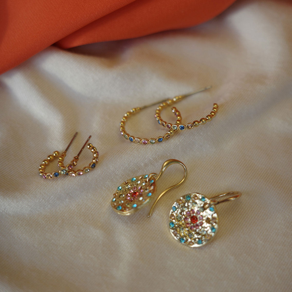 Gold earrings with colorful gemstones on a beige fabric background