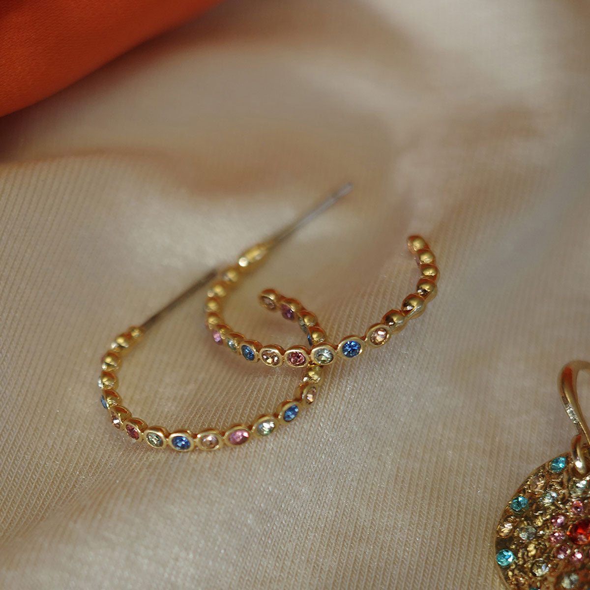 Gold hoop earrings with colorful gemstones on a beige fabric background