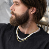 Man wearing a black shirt with two necklaces, one white and one gold, against a blurred background.