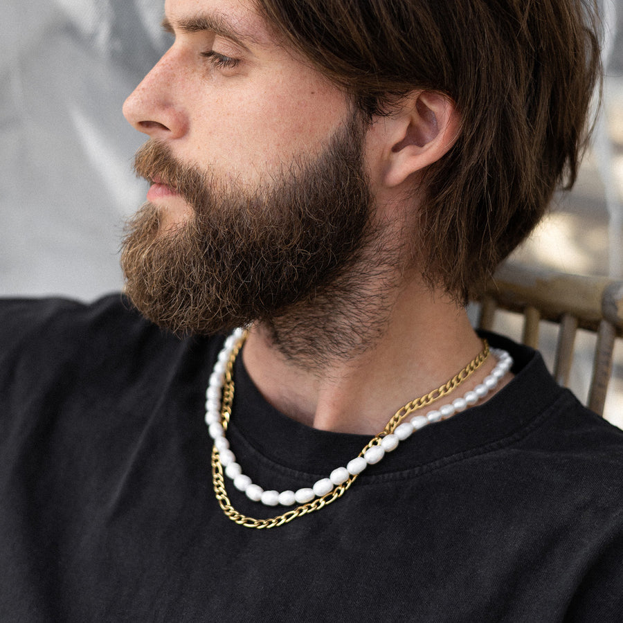 Man wearing a black shirt with two necklaces, one white and one gold, against a blurred background.