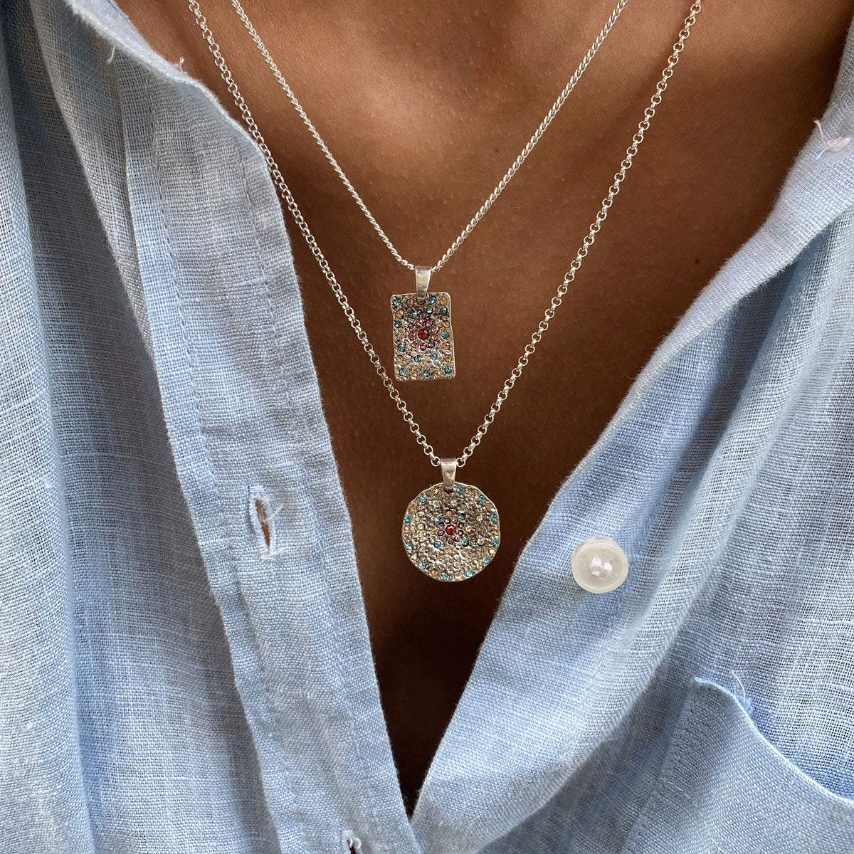 Close-up of a person wearing two silver necklaces with pendants featurning coloured crystals against a light blue shirt.