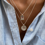 Close-up of a person wearing two silver necklaces with pendants featurning coloured crystals against a light blue shirt.