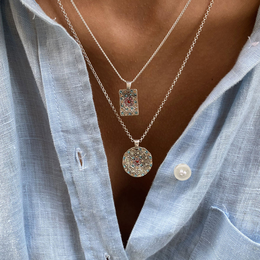 Close-up of a person wearing two silver necklaces with pendants featurning coloured crystals against a light blue shirt.