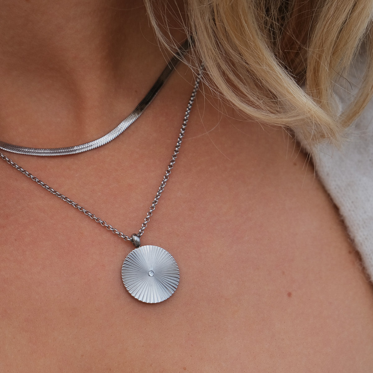 Close-up of a silver chain layered with a silver necklace with a round pendant on a person's neck.