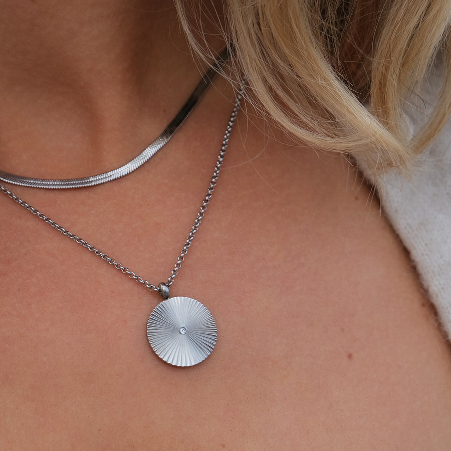 Close-up of a silver chain layered with a silver necklace with a round pendant on a person's neck.