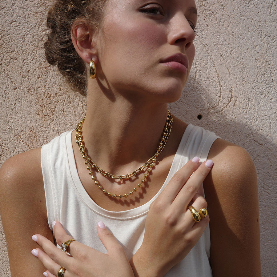 Woman wearing gold jewelry including a necklace, earrings, and rings against a beige wall.