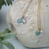 Two necklaces with blue glass and crystal pendants on a stone surface with a plant in the background