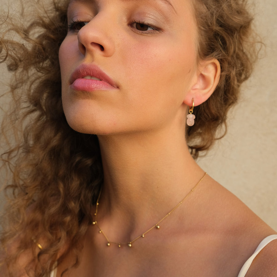 Close-up of a woman wearing a gold necklace and pink earrings against a neutral background