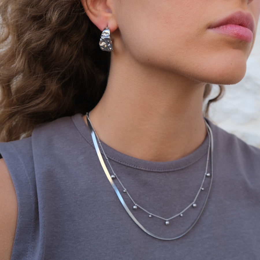 Close-up of a woman wearing a silver  ball necklace and a silver snake chain necklace  tand earrings to show layering.