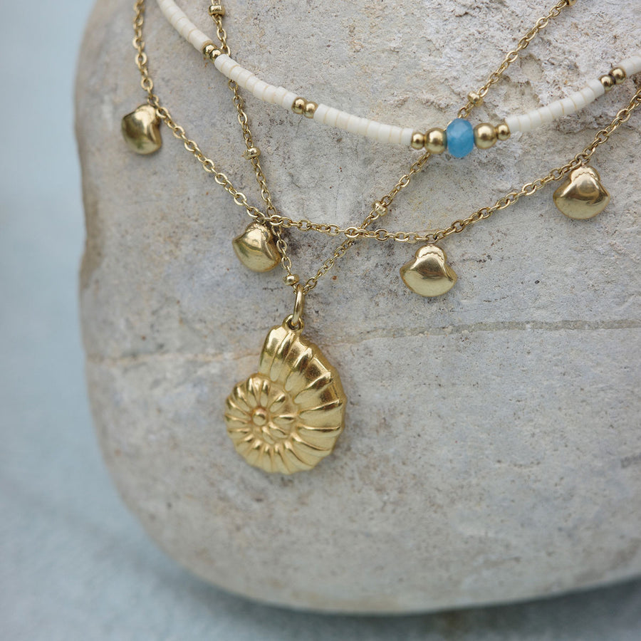 Gold necklace with shell pendant on a stone surface layered with white shell necklace with gold and blue beads
