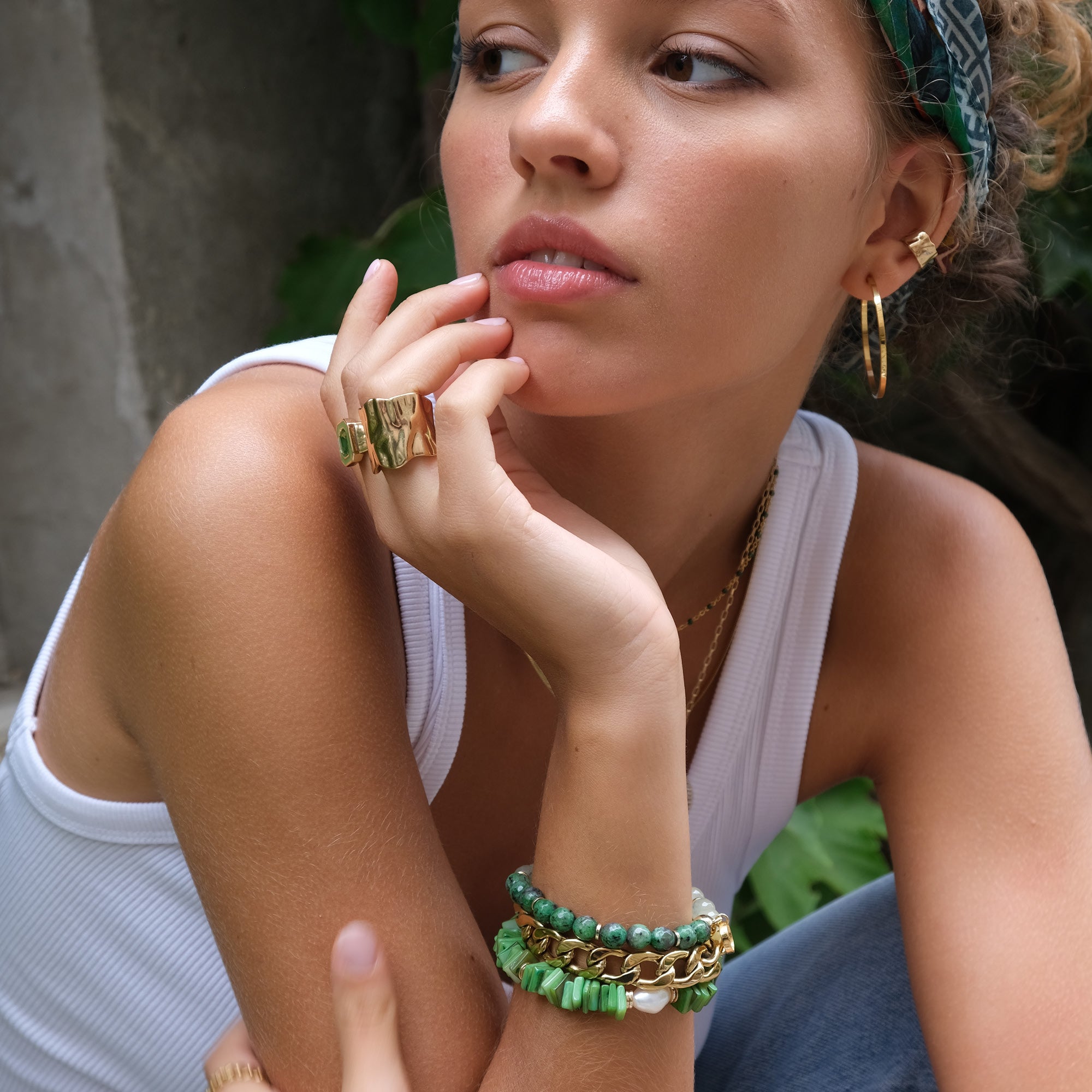 Woman wearing gold and green jewelry with a white top and headband, sitting outdoors.