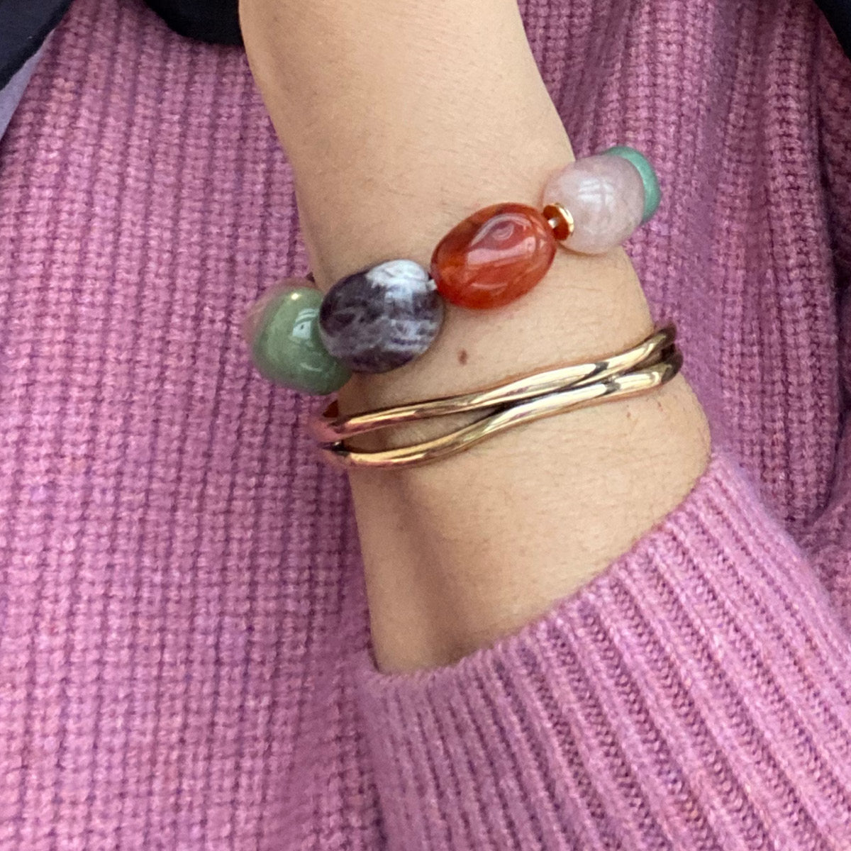 Close-up of a wrist wearing a colorful beaded bracelet and a gold bracelet, against a pink sweater background.