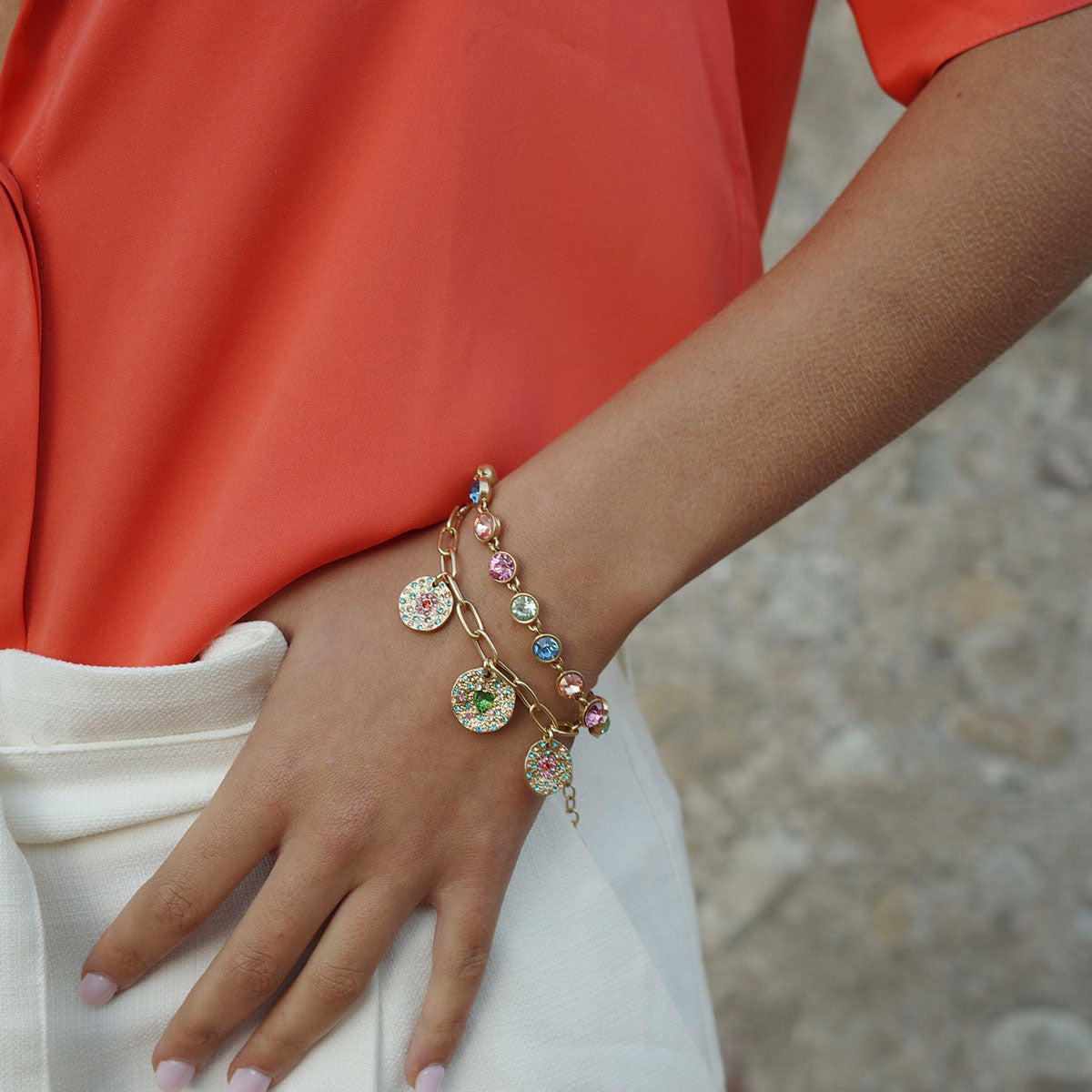 Person wearing Gold bracelet with gold charms inlaid with colourful stones and a coloured stone bracelet to show layering effect