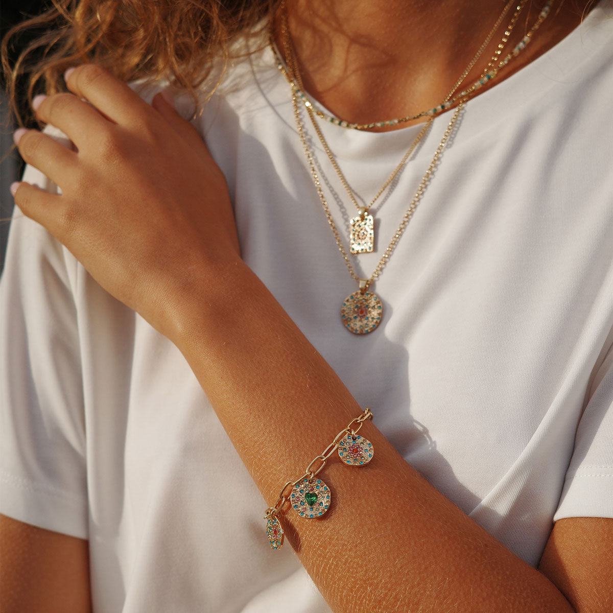Close-up of a person wearing gold necklaces and a bracelet with charms on a white shirt.