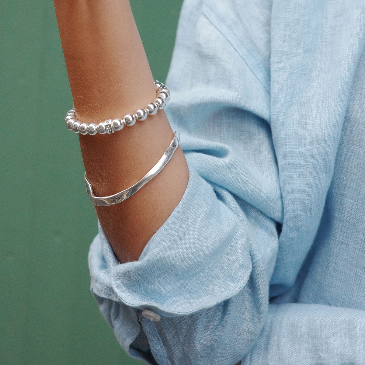 Close-up of a person's arm wearing two silver bracelets with a blurred green background