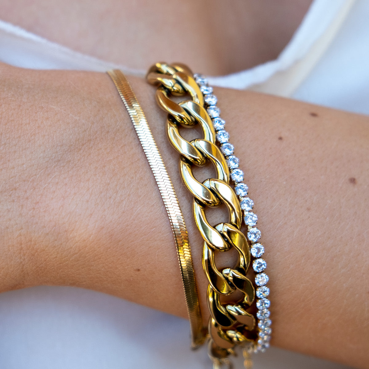 Three Gold chain bracelets, one with cruystals on a wrist against a neutral background