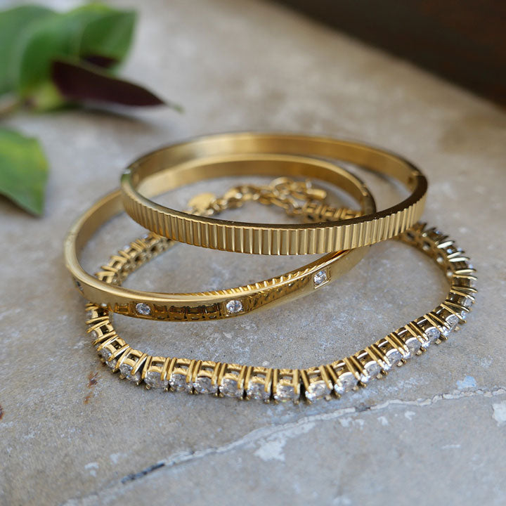Three gold bracelets with crystal accents on a stone surface.