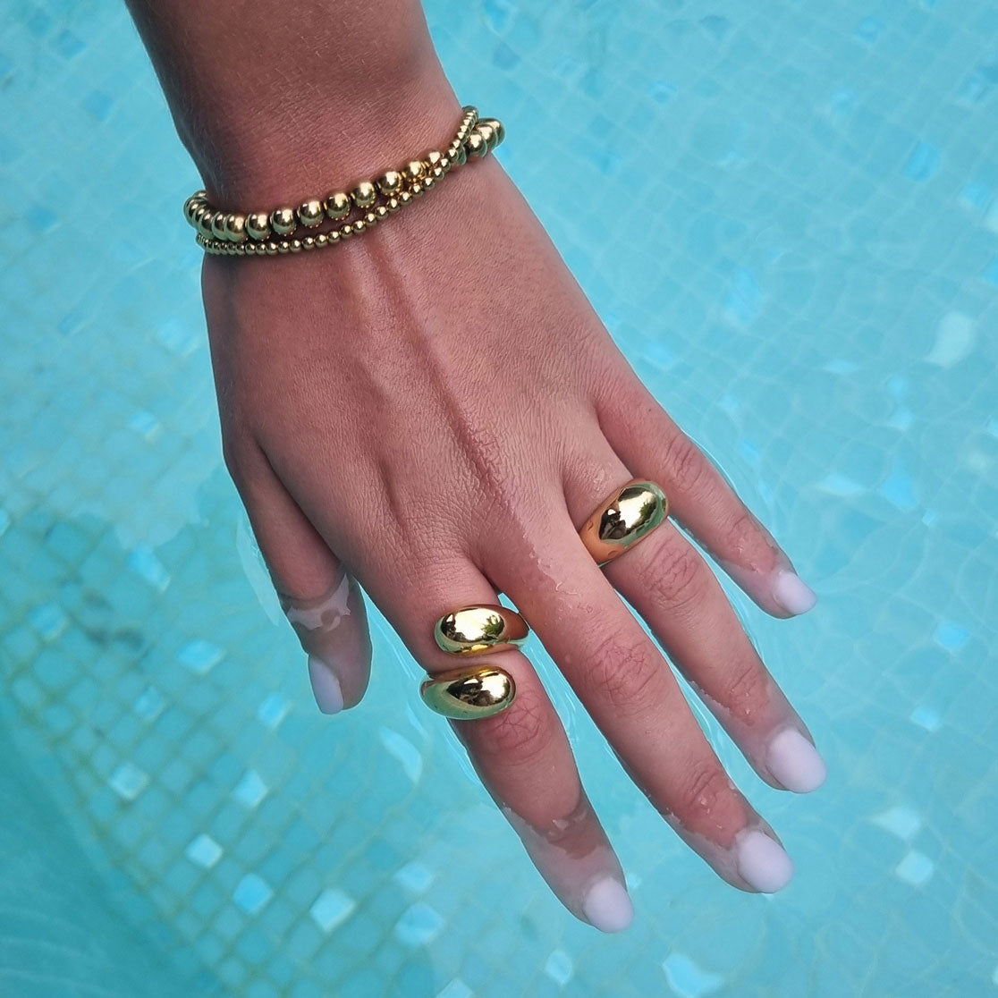 Hand with gold rings and double beaded gold bracelet against a blue tiled background