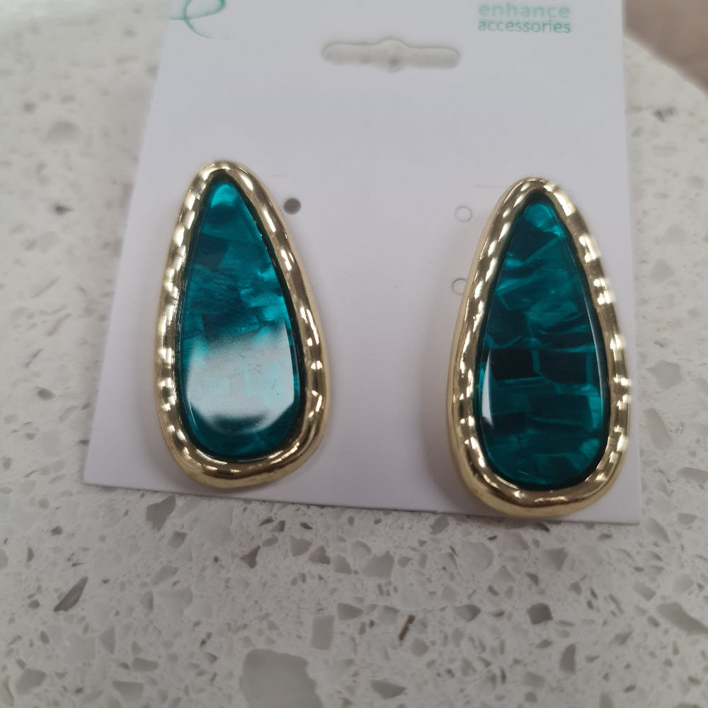 A pair of gold teardrop-shaped stud earrings with textured green-blue resin centers, displayed on a white card labeled 'enhance accessories' on a white stone surface at Cerise