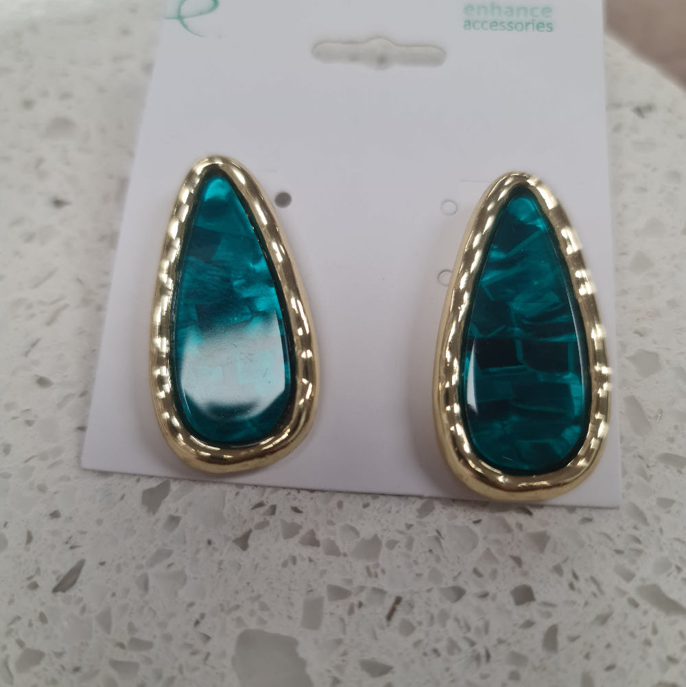 A pair of gold teardrop-shaped stud earrings with textured green-blue resin centers, displayed on a white card labeled 'enhance accessories' on a white stone surface at Cerise