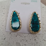 A pair of gold teardrop-shaped stud earrings with textured green-blue resin centers, displayed on a white card labeled 'enhance accessories' on a white stone surface at Cerise