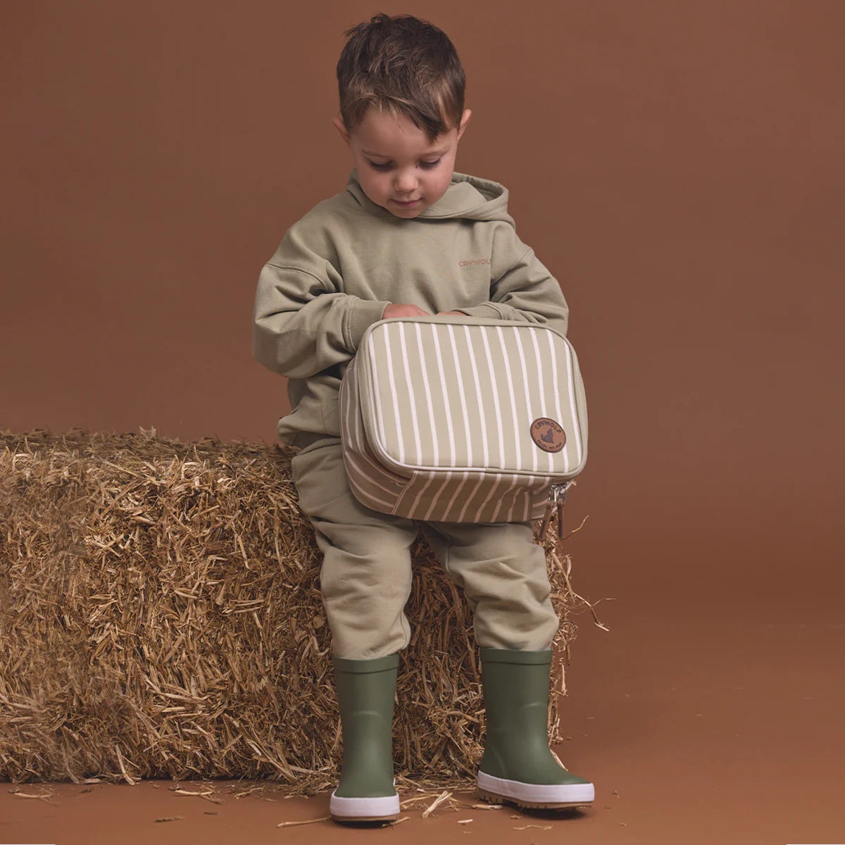 A child holding a Crywolf Everyday Lunchbag to keep kids snacks cool and luches fresh at Cerise. Made with food grade insulated lining and removeable non-toxic gel pad to keep food cool and has a handy carry handle. Available in Green Elm Stripe Design for boys and girls.