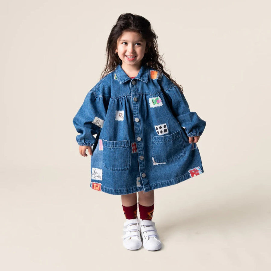 Young Girls Das Ist Maurice Denim Smock Dress with long sleeves, front patch pockets, button front, collar and gathered yoke. Has a patchwork stamp pattern on the fabric,  at Cerise NZ