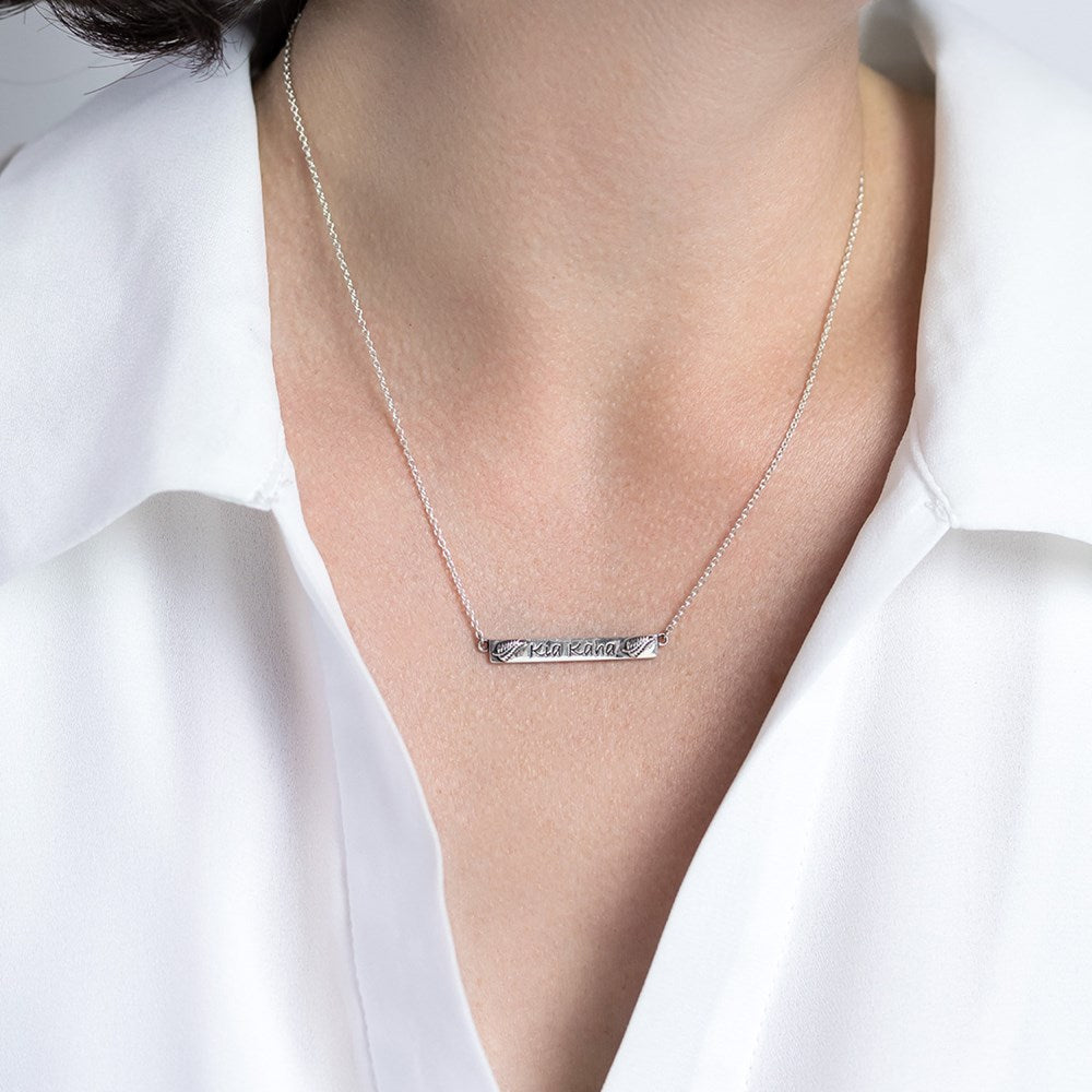 Model wearing a sterling silver necklace with a panel engraved with the words Kia Kaha and a silver fern embossed at each end of the panel.
