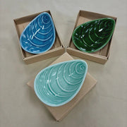 Three ceramic mussel shaped bowls in blue, green, and aqua on cardboard boxes.