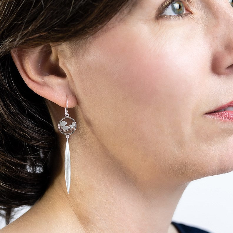 Model wearing 925 sterling silver earrings featuring two NZ Fantails also known as Piwakawaka Birds set in a circle with a flax leaf drop.