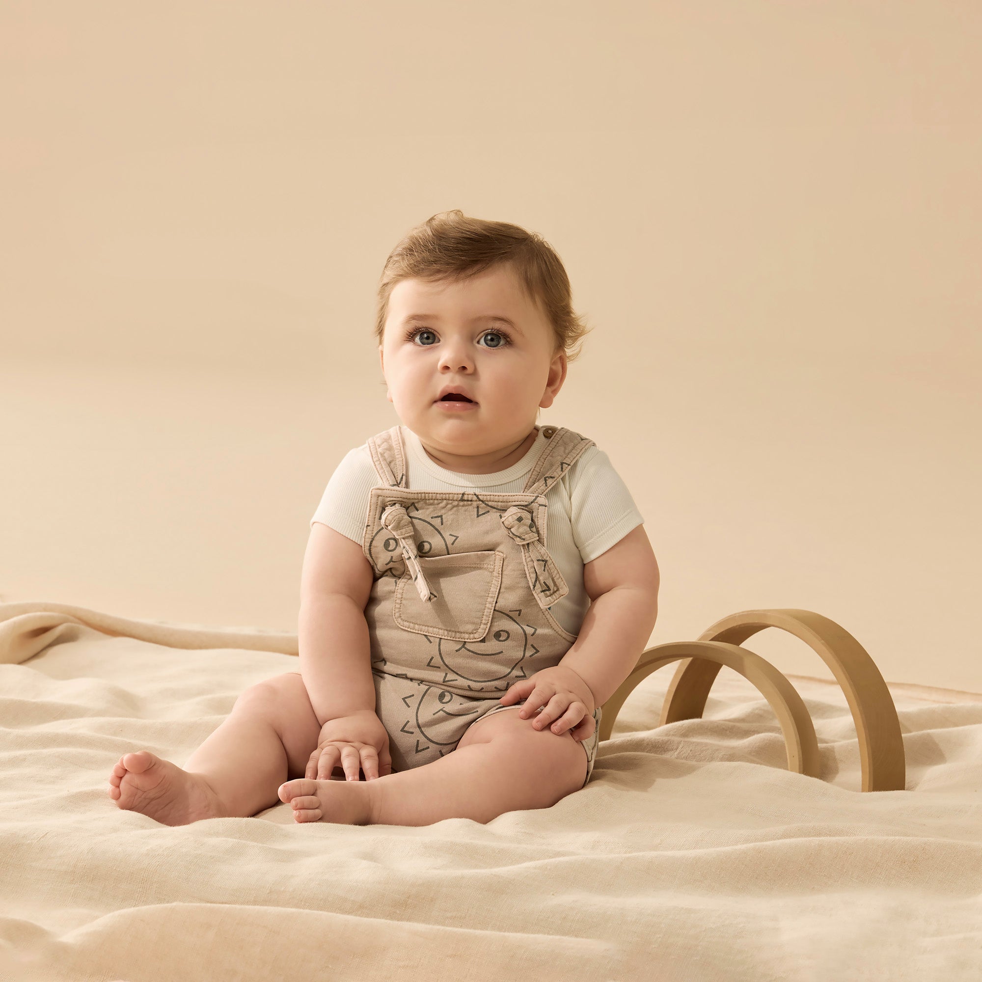Baby wearing Caramel base with brown sun faces baby overall with tie straps, front pocket and crotch dome opening.  Short legs