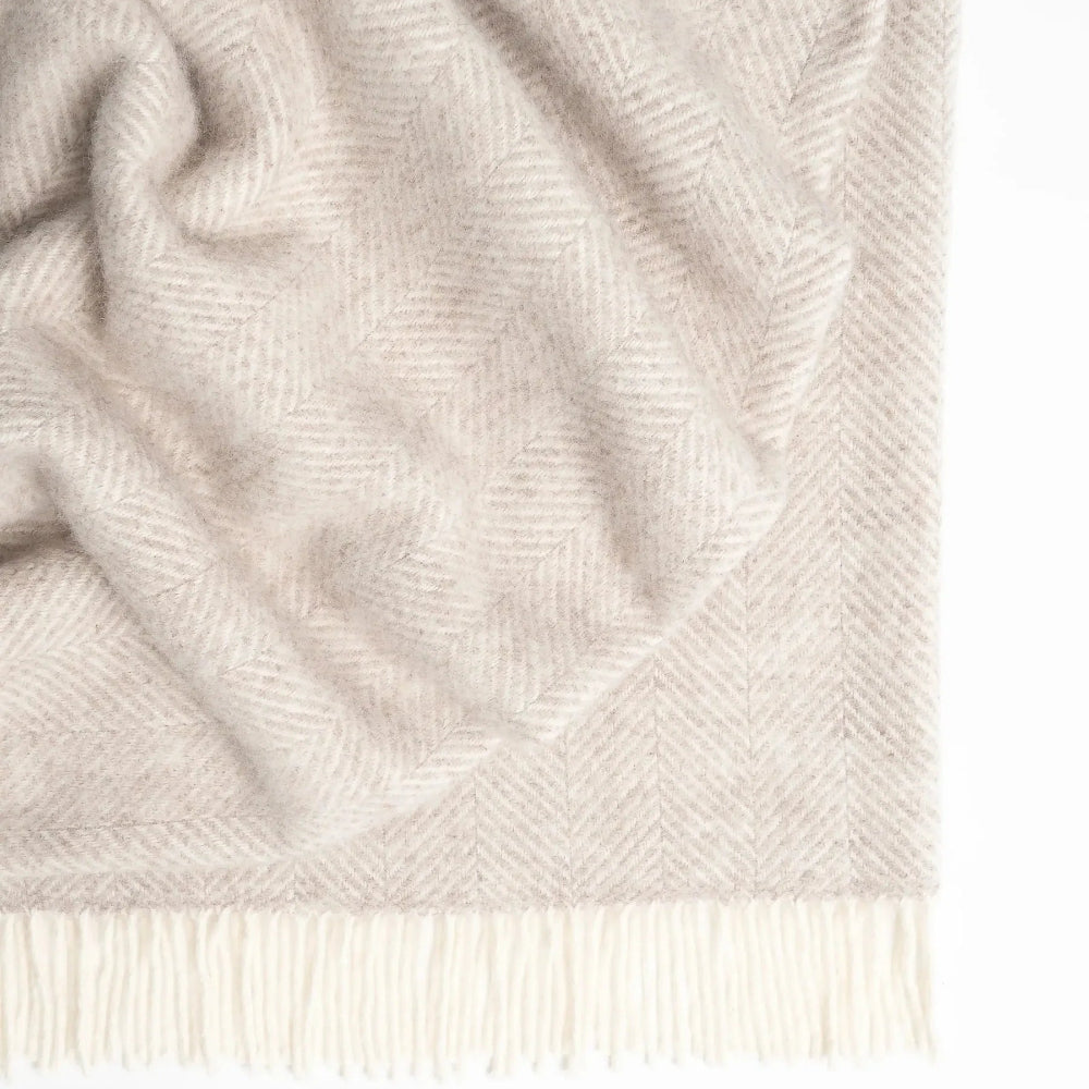 The Magnus Throw in Quartz colour at Cerise.  Made from 100% New Zealand wool, this large blanket which measures 150 x 183cm is  cosy and comfortable.  Featuring a classic herringbone weave with contrast fringe, in a natural quartz colour.