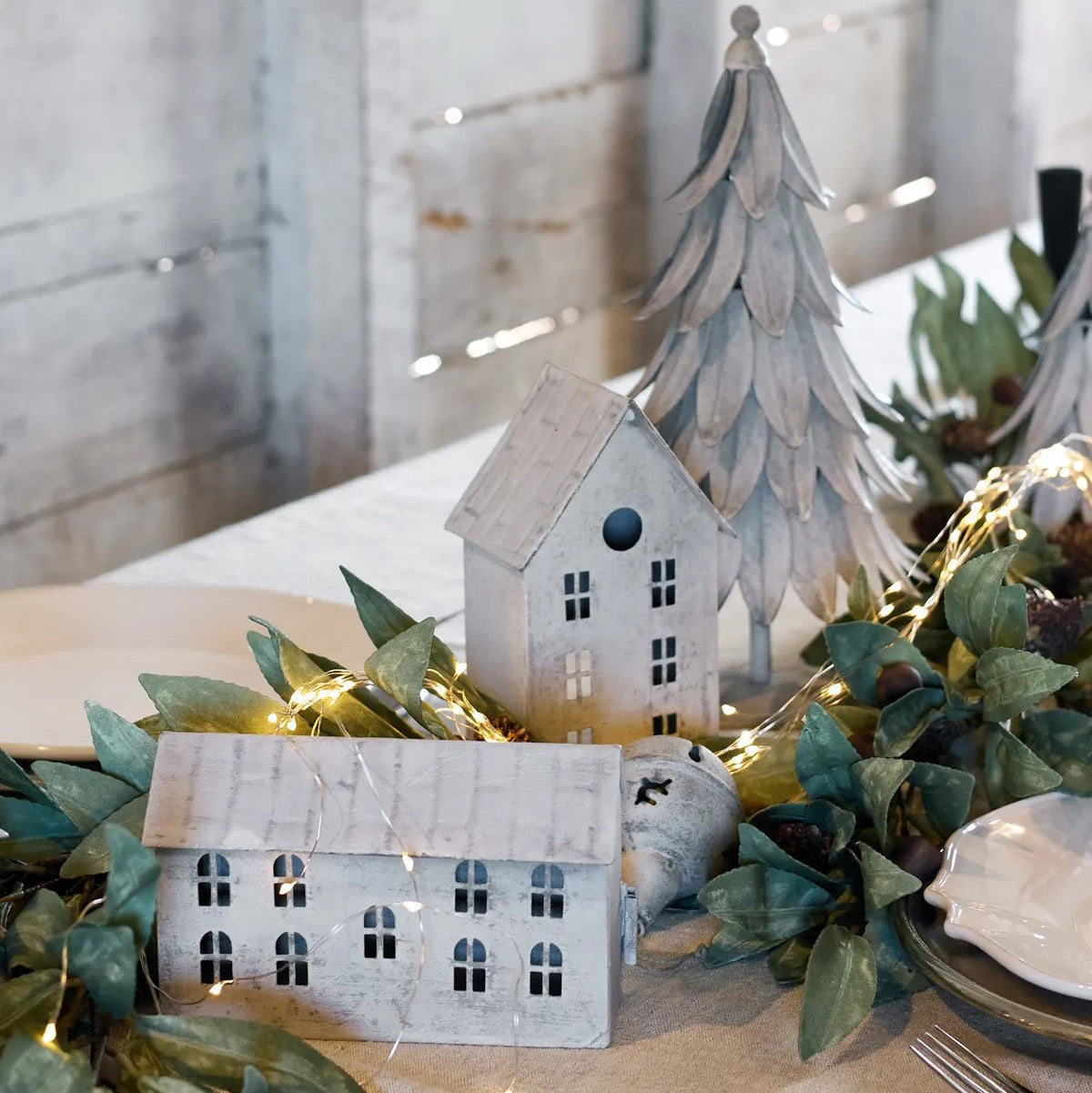 Decorative table setting with small houses, tree, and greenery on a tablecloth.