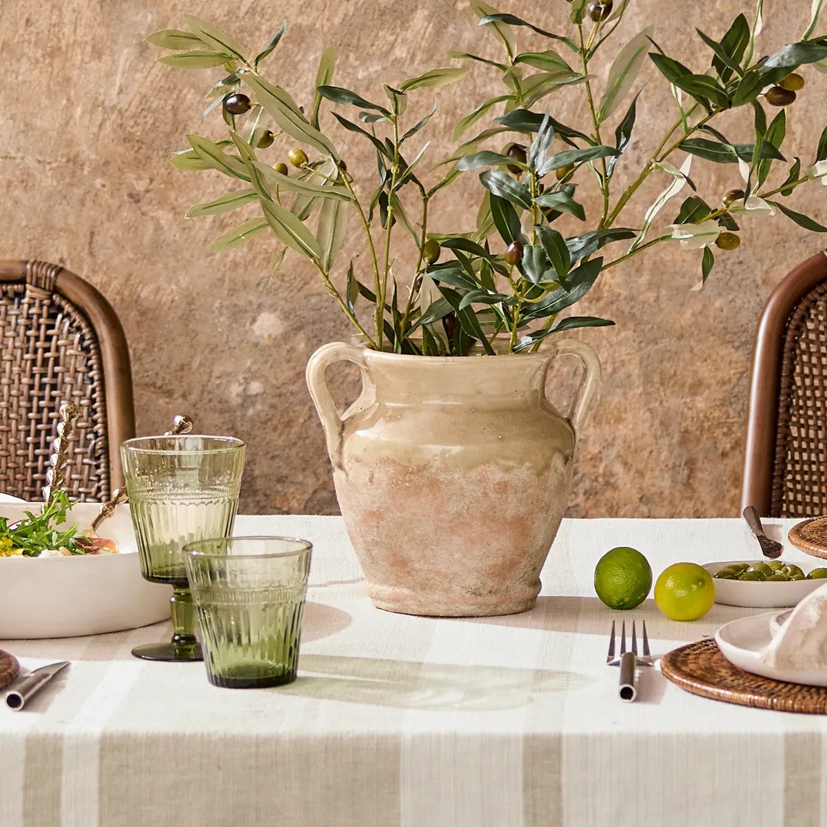 Dining table setting with a vase of greenery, glasses, and fruit on a textured wall background.
