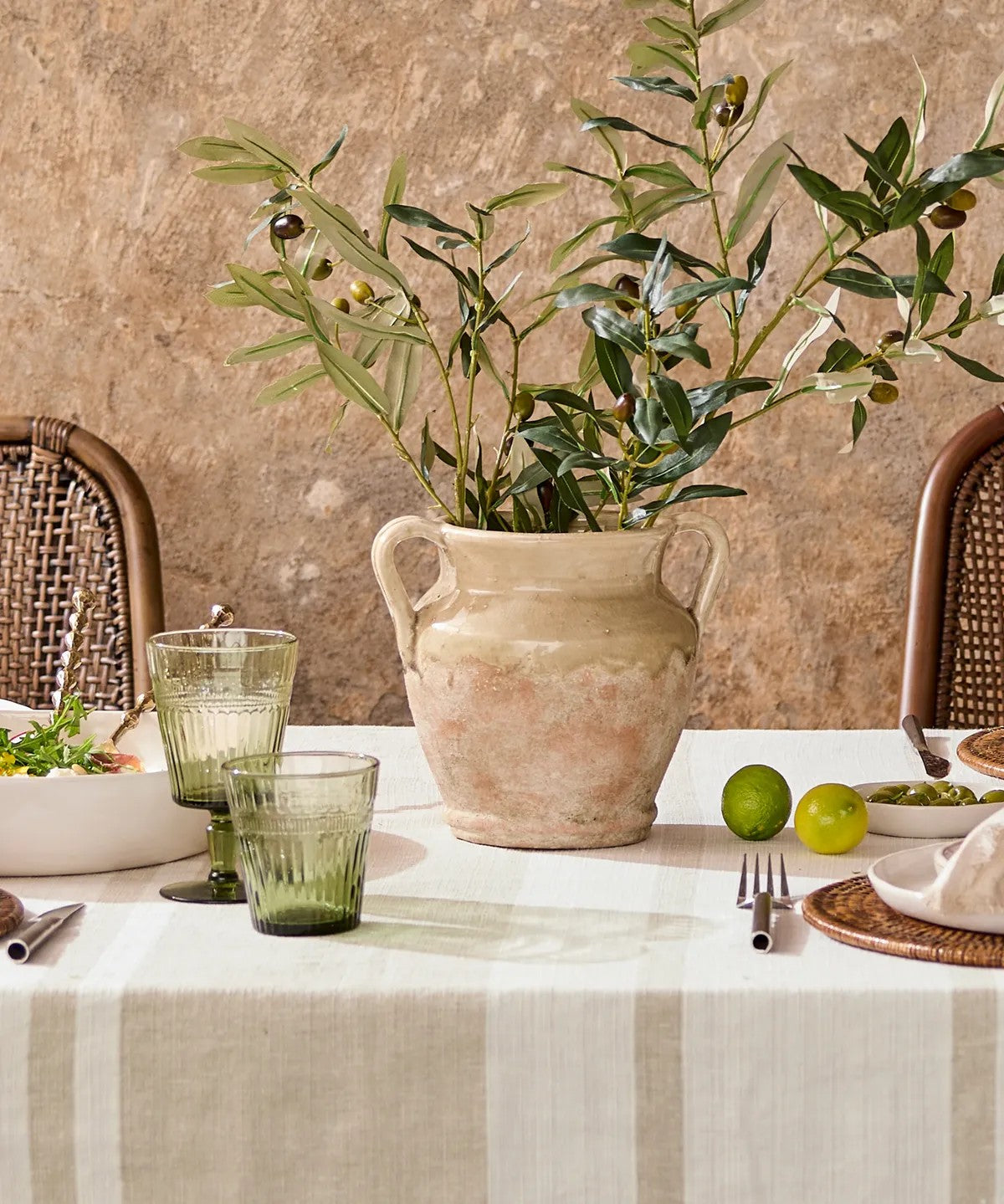 Dining table setting with a vase of greenery, glasses, and fruit on a textured wall background.