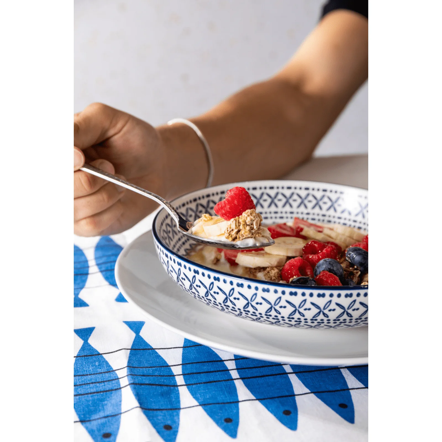 Bowl of cereal with fruit being eaten, placed on a blue and white patterned tablecloth.