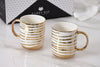 Two white mugs with gold stripes on a light surface, with 'Pampa Bay' branding in the background.
